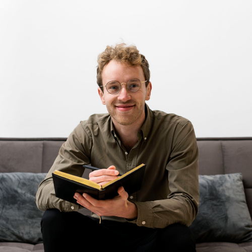 chico con lentes sonriendo sentado en un sillón con un cuaderno en la mano y una lapicera como escribiendo en él